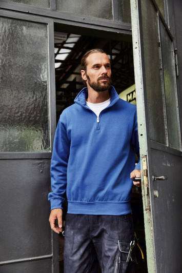 A blue half zip sweat jumper with a collar, worn by a man standing at a doorway, featuring a logo on the chest.
