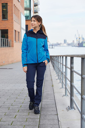 A blue and navy ladies' all-weather jacket worn by a woman walking along a waterfront promenade.