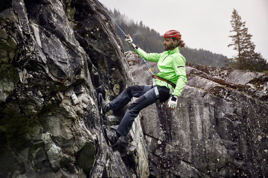 A man in a bright green jacket and climbing gear descends a rocky cliff, wearing black zip-off trekking pants.