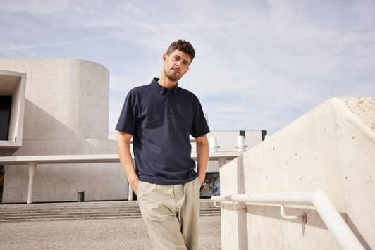A man wearing a navy plain polo shirt stands casually against a backdrop of modern architecture.