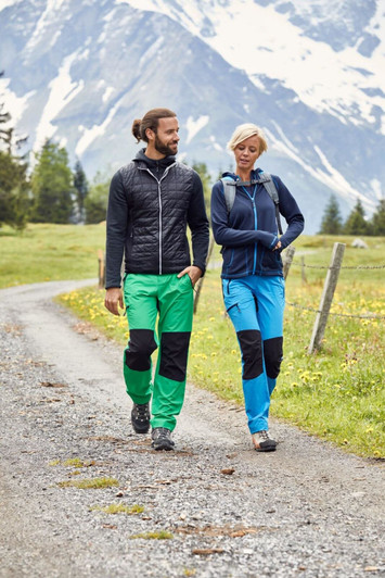 Two people walking on a trail, wearing green and blue trekking pants with black patches, set against a mountainous background.
