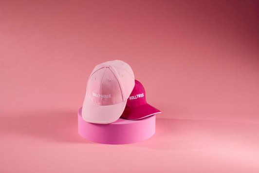 A pink and a magenta 6 Panel Sandwich Cap are displayed against a pink background, with a logo on each cap.