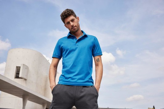 A man wearing a blue mercerised polo shirt stands outdoors, hands in pockets, against a cloudy sky backdrop.