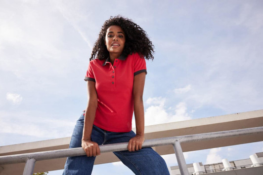 A woman in a red polo shirt poses confidently while leaning on a railing, under a clear sky. The shirt has a logo.