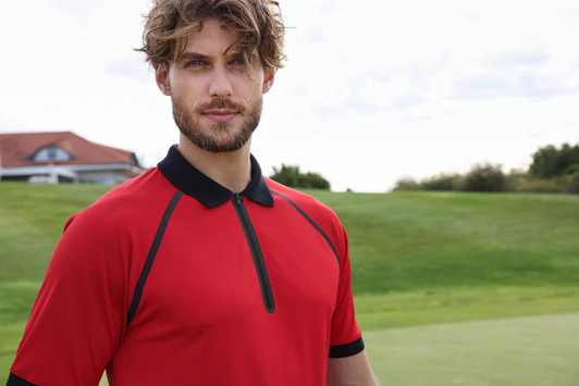 A man in a red zip polo shirt with black accents stands on a golf course background. The shirt features a collar and a logo.