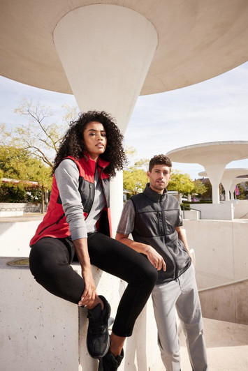 A ladies' fleece vest in red, paired with a grey vest, worn by models in a modern architectural setting.