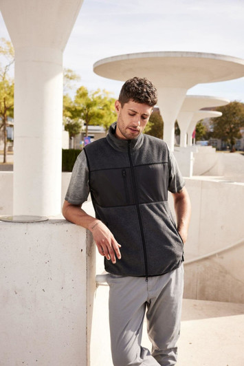 A men's fleece vest in black, featuring a zip front and a logo, worn by a model in an outdoor setting.