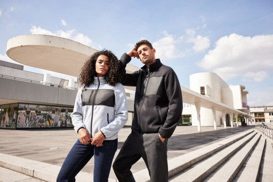 A fleece jacket in light blue and black, worn by two models outside a modern building, featuring a logo.