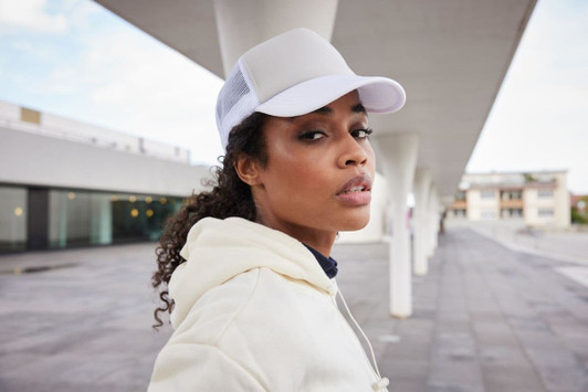 A woman wearing a white 5 Panel Polyester Mesh Trucker cap, with a light-coloured hoodie, standing against a modern backdrop.