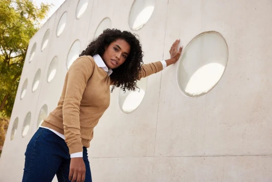 A knitted jumper in tan with a white collar, worn by a woman against a textured wall with circular cutouts.