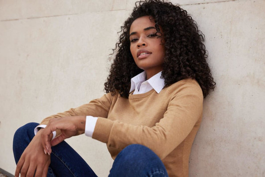 A knitted round-neck pullover in tan, featuring a woman sitting against a wall, wearing a collared shirt underneath.