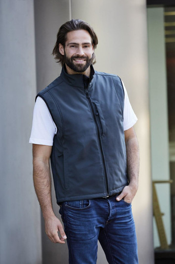 A men's softshell vest in dark grey, featuring a front zip and a logo. Worn over a white t-shirt and blue jeans.