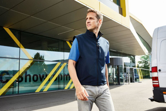 A man wearing a navy blue softshell vest with blue sleeves, standing outside a modern building.