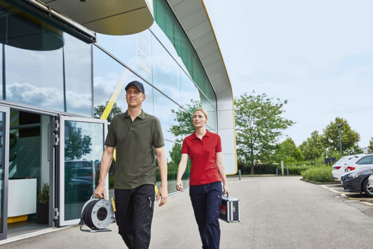 A man in a green polo shirt and a woman in a red polo shirt carry equipment outside a modern building. Both shirts have logos.