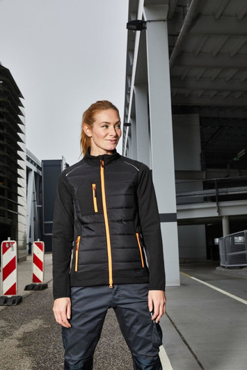 A black hybrid jacket featuring orange accents, with zippered pockets, set against an industrial background.
