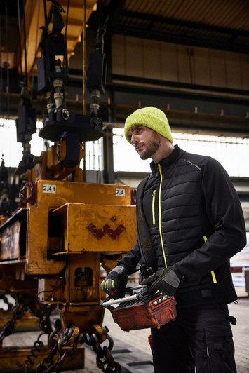 A man wearing a black and yellow hybrid jacket stands in an industrial setting, holding a remote control.