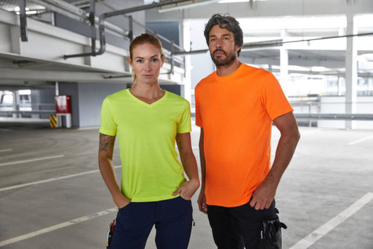 Two individuals stand together in a parking garage, wearing a neon yellow and an orange T-shirt. The shirts have a logo.