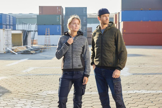 A padded hybrid jacket in grey and black, worn by a woman and a man outdoors near shipping containers. The jackets feature a logo.