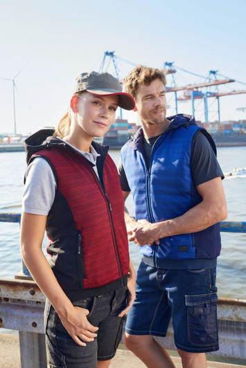 A red and black padded hybrid vest is worn by a woman, alongside a man in a blue vest, both by a port.