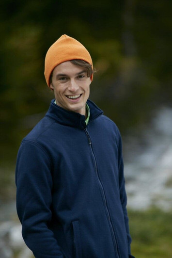 A man wearing an orange beanie and a navy blue jacket, smiling outdoors with a blurred natural background.