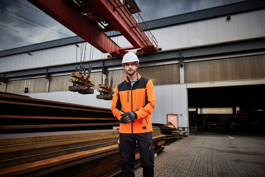 A man in an orange workwear jacket stands outside a warehouse, wearing gloves and a hard hat. The setting includes steel beams and a crane.