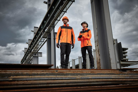 Two individuals wearing bright orange work jackets stand on metal beams at a construction site, with overcast skies in the background.