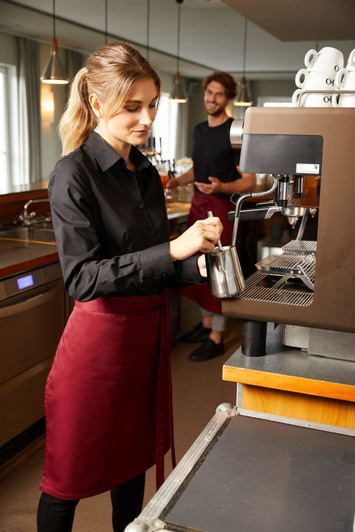 A woman wearing a black shirt and a maroon apron prepares coffee at a café, with a barista in the background.