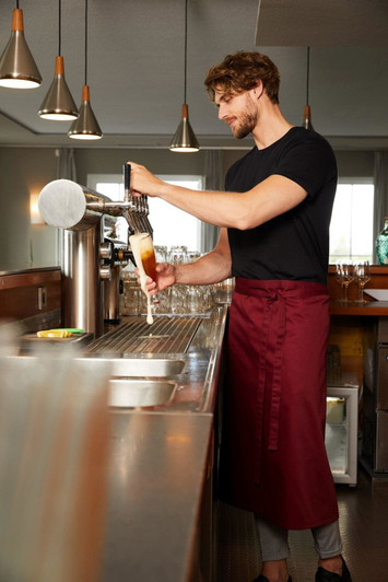 A man wearing a burgundy apron pours a drink from a tap in a bar setting. The image features modern pendant lights.