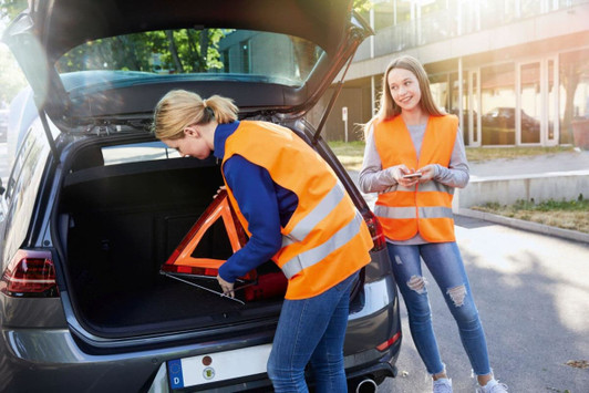 Two individuals wearing bright orange safety vests work near a car, with one placing a warning triangle in the boot.