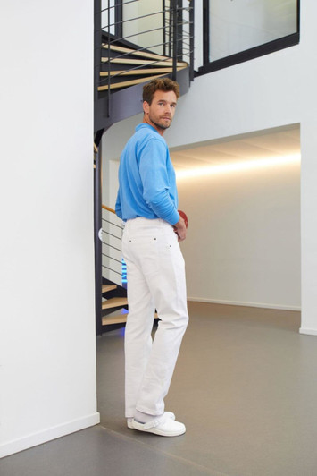 A man in a blue shirt wears white stretch pants, standing near a staircase in a modern interior. The pants have a logo.