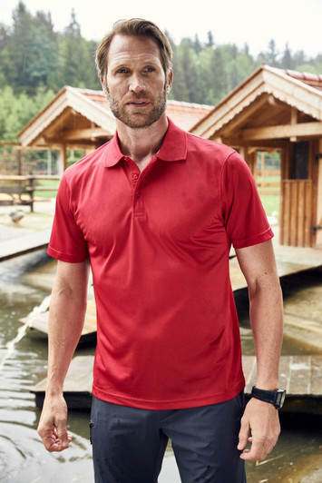A red men's polo shirt is displayed on a man standing near wooden structures by water. The shirt features a logo.