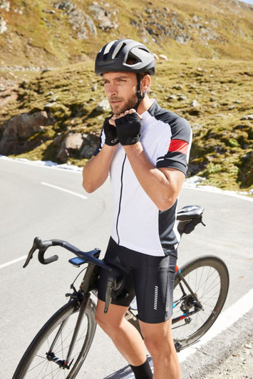 A man in a black and white cycling jersey adjusts his gloves while standing next to a bicycle on a mountain road.