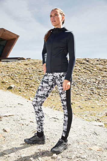A black zip hoodie worn with patterned leggings, set against a natural outdoor background. The model is standing confidently.