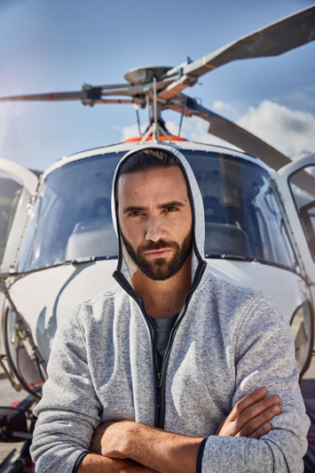 A men's knitted fleece hoodie in grey, worn by a man standing in front of a helicopter, featuring a hood and branding.