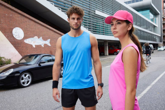 A blue men's active tank top and a pink women's tank top are modelled in an urban setting. Both feature logos.
