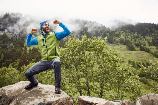 A man wearing a green and blue knitted fleece vest poses on a rock outdoors, surrounded by greenery and mountains.