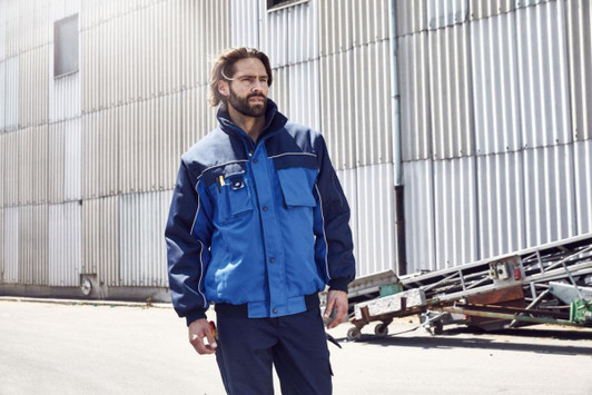 A workwear jacket in navy and blue, featuring a front pocket and logo, worn by a man in an industrial setting.