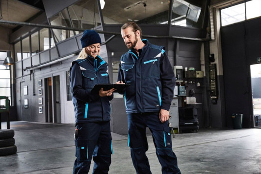 A padded work vest in navy blue with teal accents, worn by two people in a workshop setting, one with a clipboard.