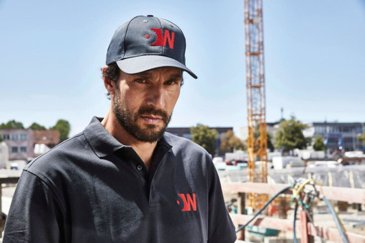 A man wears a black polo shirt and matching cap, featuring a logo, against a construction site backdrop.