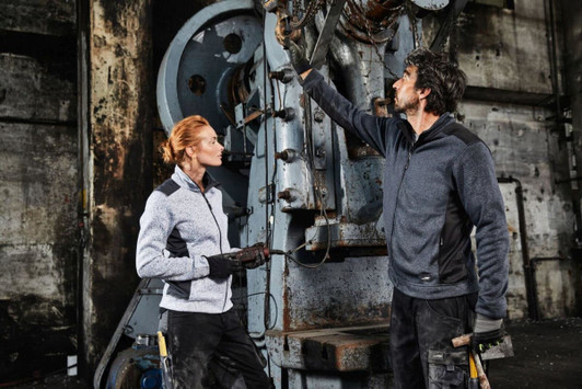 A grey fleece jacket worn by a woman, paired with a male model in a darker fleece jacket, both in an industrial setting.