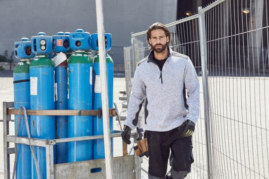 A man wearing a grey knitted fleece half-zip top stands near blue gas cylinders, with a worksite backdrop.