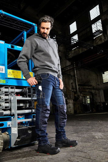 A man wearing grey workwear and blue stretch jeans stands near a worksite with industrial equipment.