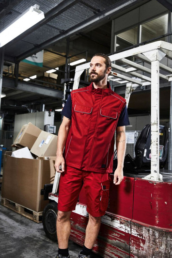 A man wearing a red softshell light vest and matching shorts stands in an industrial setting, with boxes in the background.