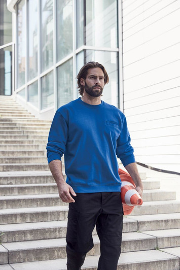 A man wearing a blue round sweat pocket jumper walks down stairs, holding an orange and white item. The setting is modern and outdoor.