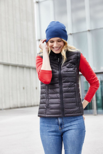 A woman wears a black puffer vest over a red shirt and a blue beanie, smiling outdoors.