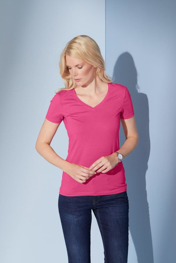 A fitted ladies' t-shirt in pink with a V-neck design, paired with dark blue jeans. The model stands against a light blue background.