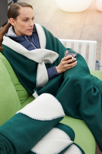 A green bonded fleece blanket with a white trim draped over a woman sitting on a couch, holding a drink.