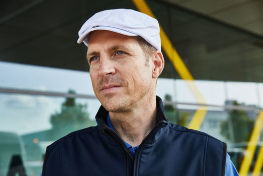 A man wearing a white Cabrio Cap and a dark vest, looking away against a modern architectural background.