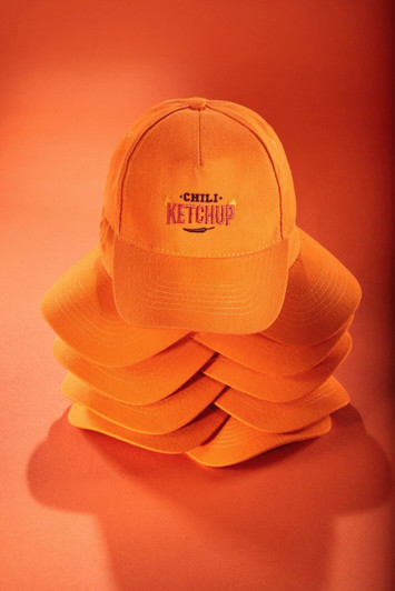 An orange 5-panel cap is displayed on top of a stack of similar caps, featuring a logo on the front.