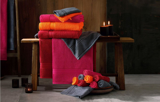 A stack of vibrant towels in pink, orange, and grey arranged on a wooden bench, with candles nearby.